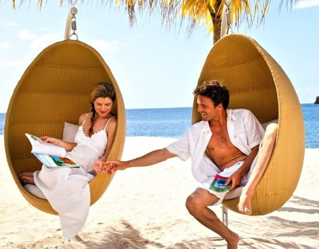 A couple relaxing in hanging chairs on a sandy beach, holding hands with the ocean in the background.