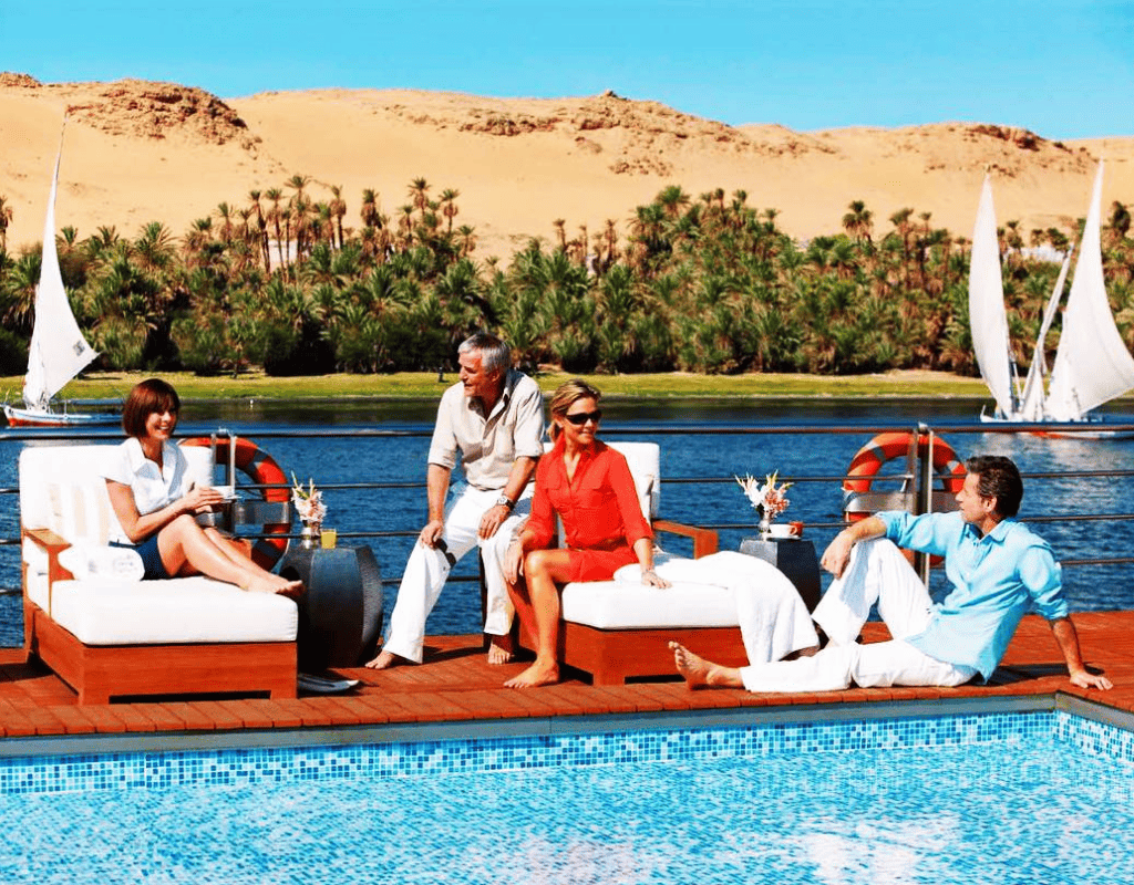 A group of friends relaxing by the pool on a Nile River cruise, with feluccas sailing in the background and golden desert dunes.